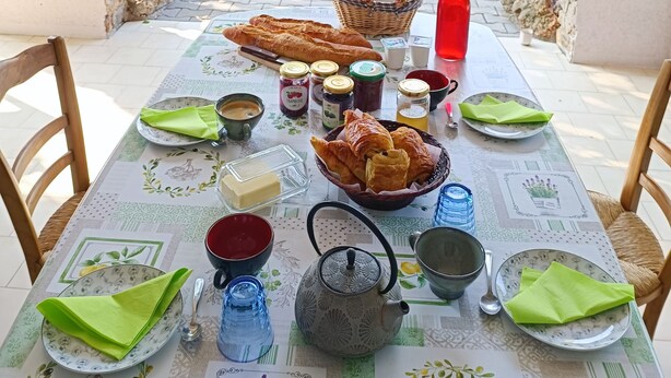 Ein Frühstückstisch mit Brot, Gebäck, Beilagen und Getränken mit Blick auf einen üppigen Garten und Berge.
