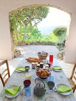 Una mesa lista para el desayuno, con pan, bollería, salsas y bebidas, con vistas a un jardín exuberante y montañas.