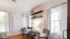A room with hardwood flooring, a grey upholstered armchair, a small table with a lamp, and a shelf with decorative items. - Modern luxury with classic Fredricksburg charm just blocks from Main Street (Fredericksburg)