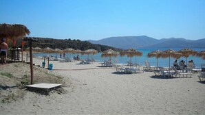 Ein Strand mit Liegestühlen und Sonnenschirmen, einer Holzkonstruktion sowie Blick auf Meer und Berge.