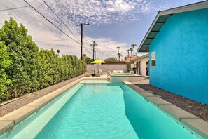 A rectangular swimming pool with a light blue interior, surrounded by a low stone wall and a hedge. There is a yellow umbrella and outdoor furniture in the background.