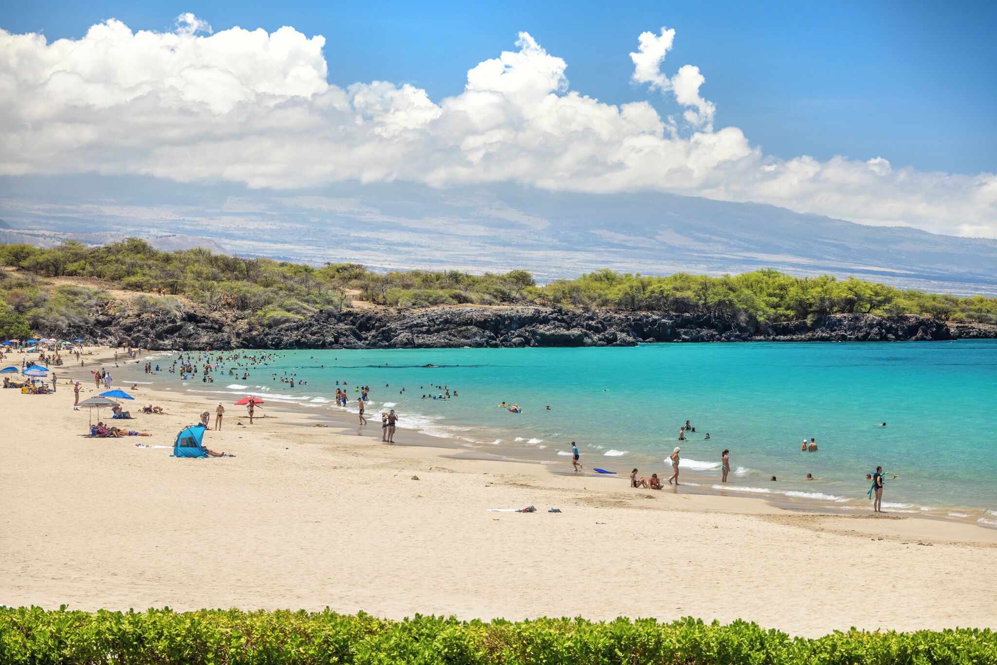 A beach with clear turquoise water, people swimming and sunbathing, and a rocky shoreline.