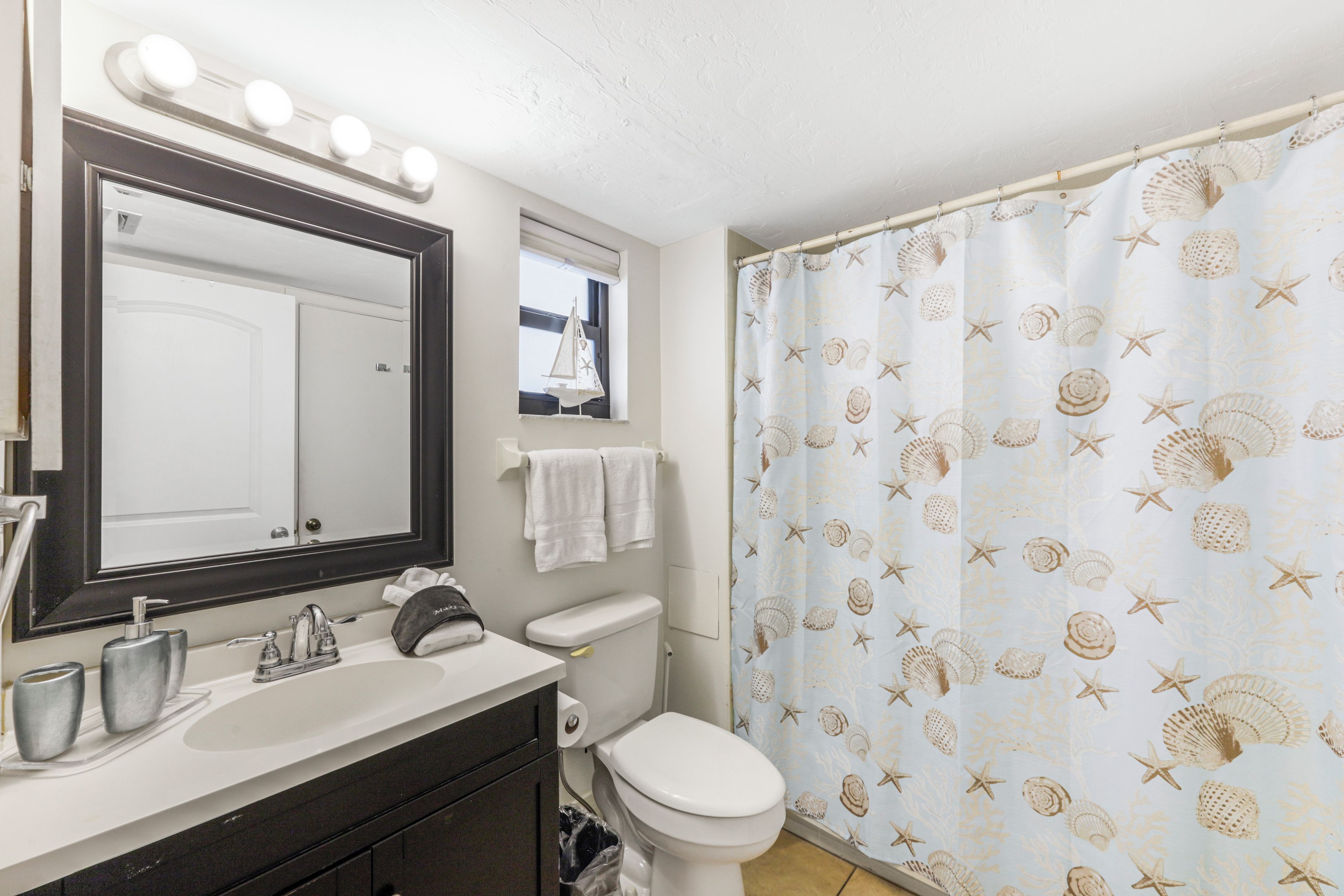 Un baño con lavamanos blanco, un espejo con iluminación, inodoro y cortina de ducha con estampado de conchas marinas.