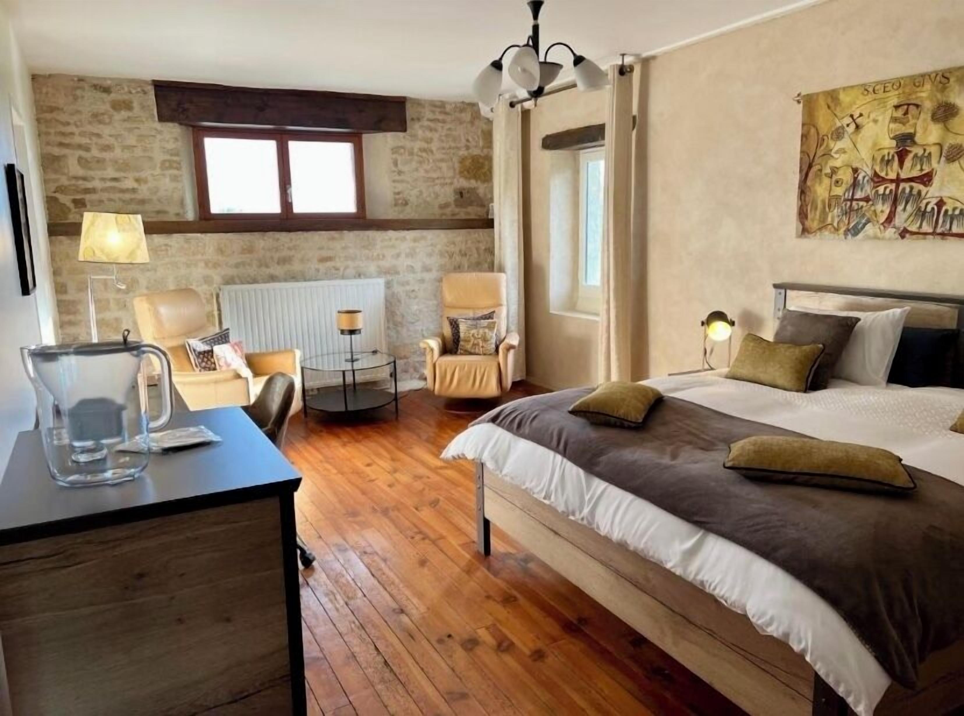 A bedroom with a bed, two chairs, a table, a radiator, a window, and a painting on the wall.