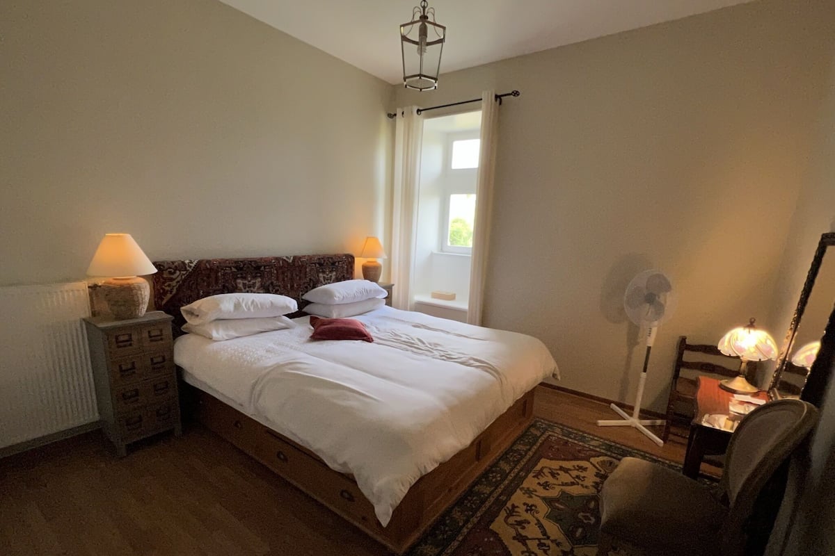 Een opgemaakt bed met witte lakens en kussens, een houten hoofdbord, een nachtkastje met een lamp, een vloerventilator en een stoel.