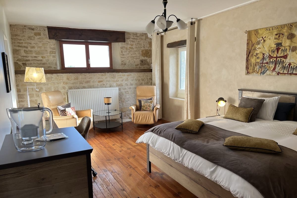 Een slaapkamer met een bed, een stoel, een tafel met een karaf, een radiator en een raam.