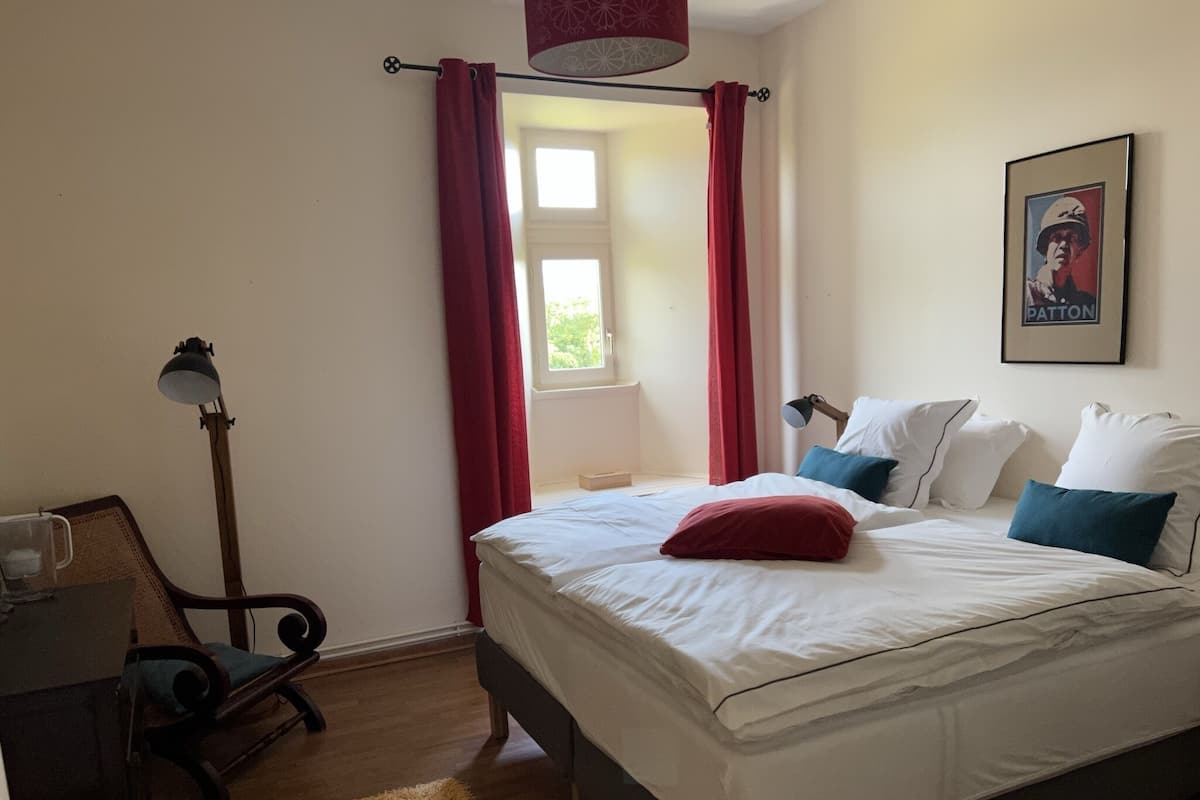 Een slaapkamer met een bed, kussens, een lamp, een stoel en een raam met gordijnen.