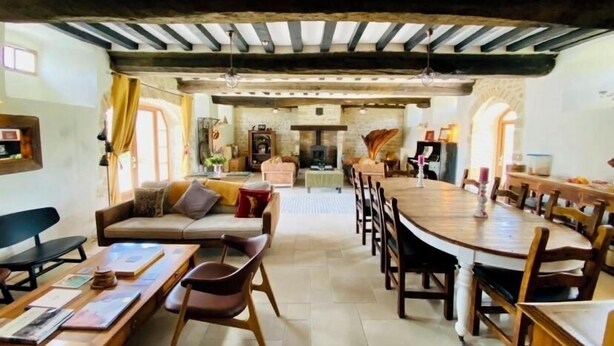 A spacious room with a high ceiling, exposed beams, a fireplace, a dining area with a long table and chairs, and a cozy seating area with a sofa and armchairs.