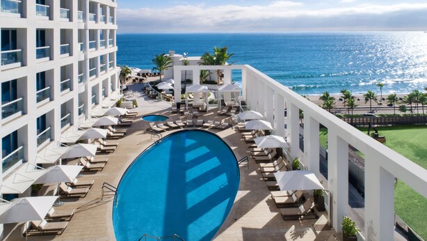 A hotel pool area with a view of the ocean, lounge chairs, umbrellas, and a large swimming pool.