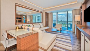 A modern hotel room with a glass desk, sofa, and armchair, large windows with a city view, and a flat-screen TV.