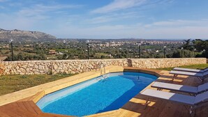 Une piscine bordée d’un mur en pierre et dotée d’une terrasse en bois, entourée de chaises longues, et offrant une vue sur une ville et des montagnes en arrière-plan.