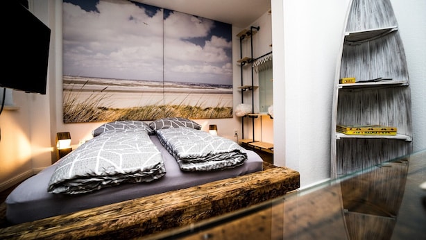 A bedroom with a bed, a TV, a wall-mounted shelf with books, and a beach view mural. - Wismarer-heckflosse (Wismar)