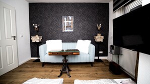 A room with a light blue sofa, a small wooden table, two wall-mounted lamps, a television, and a radiator. - Wismarer-heckflosse (Wismar)