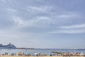 Uma praia com um navio de cruzeiro atracado, areal, guarda-sóis e pessoas.