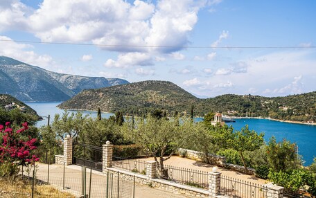 A coastal landscape with a clear view of the sea, mountains, and a fenced property with a stone wall.