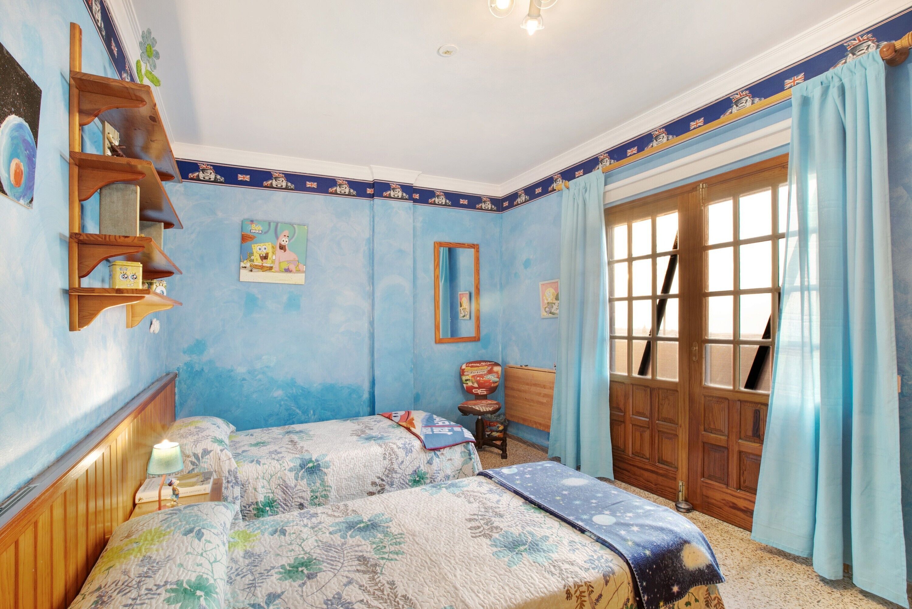 A bedroom with two beds, a wooden shelf, a chair, a window with curtains, and a door.