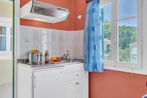 Une cuisine avec des meubles blancs, une hotte de cuisinière en acier inoxydable, une fenêtre ornée d’un rideau bleu et une vue sur la verdure à l’extérieur.