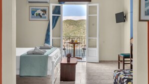 Une chambre avec un lit, une petite table et une chaise, un balcon donnant sur un paysage vallonné, et un téléviseur fixé au mur.