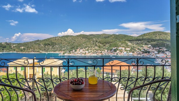 Un balcon avec une table, un saladier de fruits et une carafe de boisson, donnant sur une ville côtière avec ses maisons et sa plage.