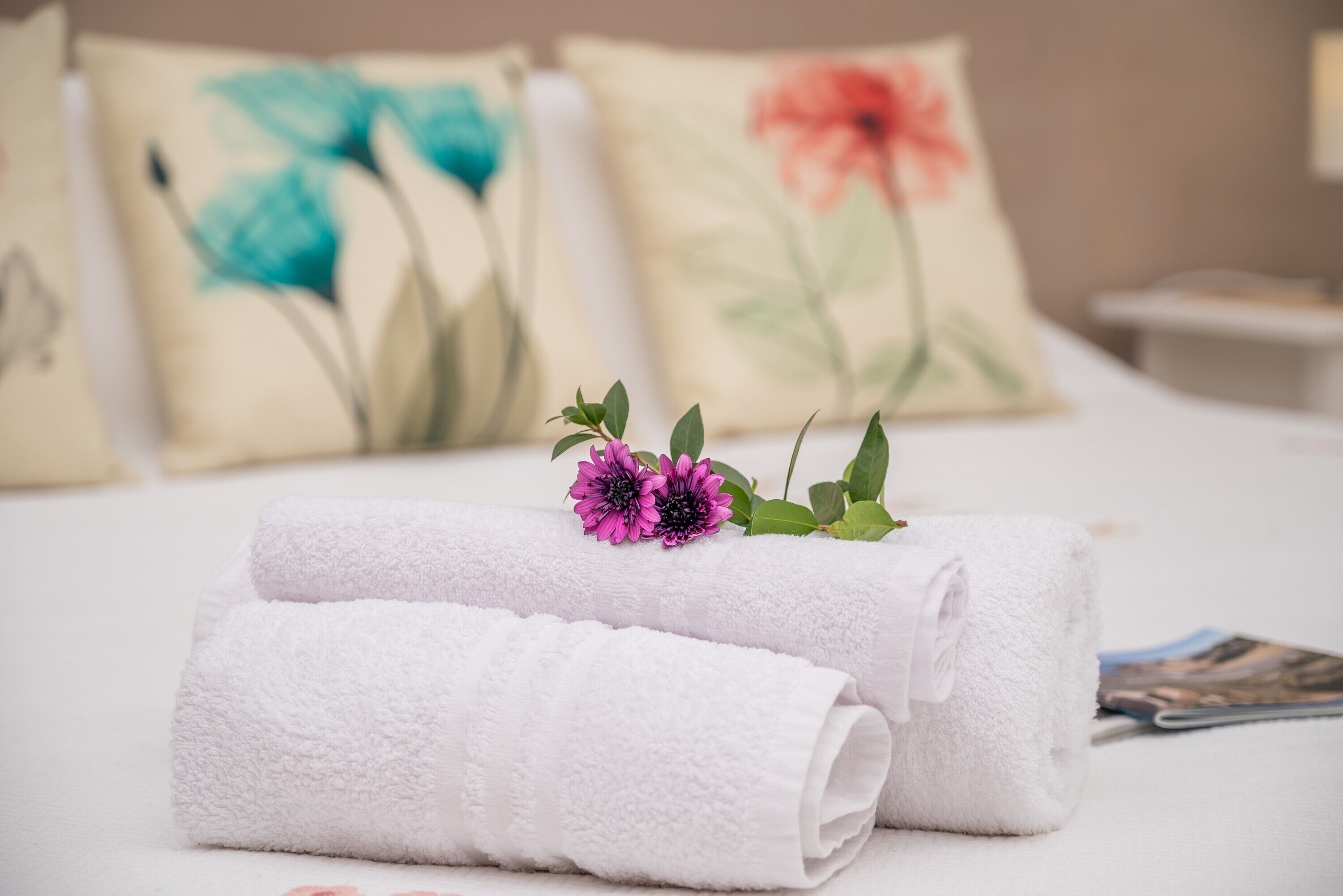 Ein ordentlich gemachtes Bett mit aufgerollten weißen Handtüchern und einem dekorativen Kissen mit floralem Muster.