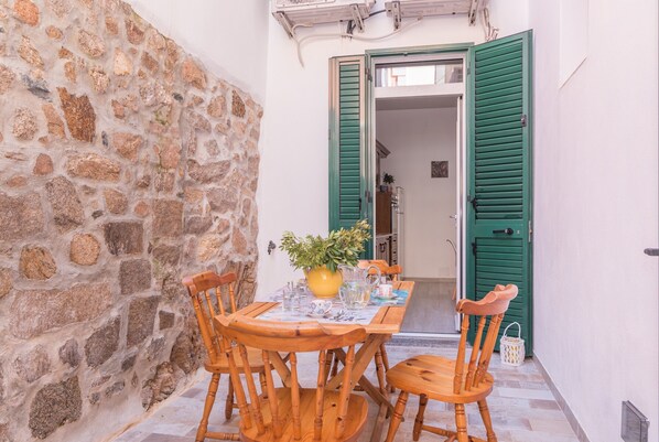 Un mur de pierre, une table en bois avec des chaises, une plante en pot et une porte avec des volets verts.