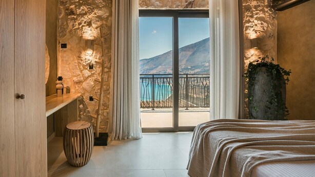A bedroom with a stone wall, a wooden bed, a small round stool, and a view of the sea through a balcony. - Adrias Villa (Kefalonia)
