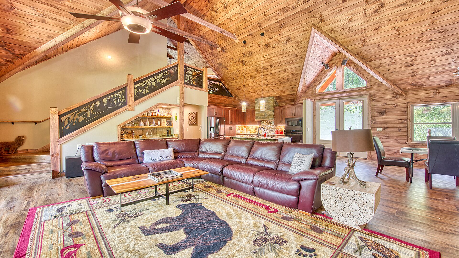 A spacious living room with a large leather sectional sofa, a wooden staircase, and a kitchen area.