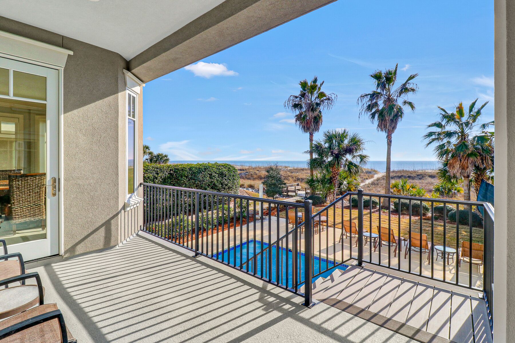 A balcony with a view of a pool and palm trees, a clear sky, and a distant beach.