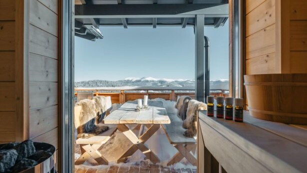 A wooden cabin with a dining area featuring a table and benches, a fireplace, and a view of snow-covered mountains.
