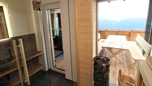 A wooden cabin with a glass door leading to a balcony with a table and chairs, a stone fireplace, and a wooden ladder.