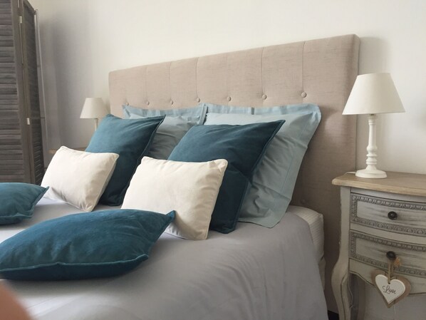 A bed with a tufted headboard, multiple pillows in shades of blue and white, a bedside table with a lamp, and a drawer with a heart-shaped decoration.