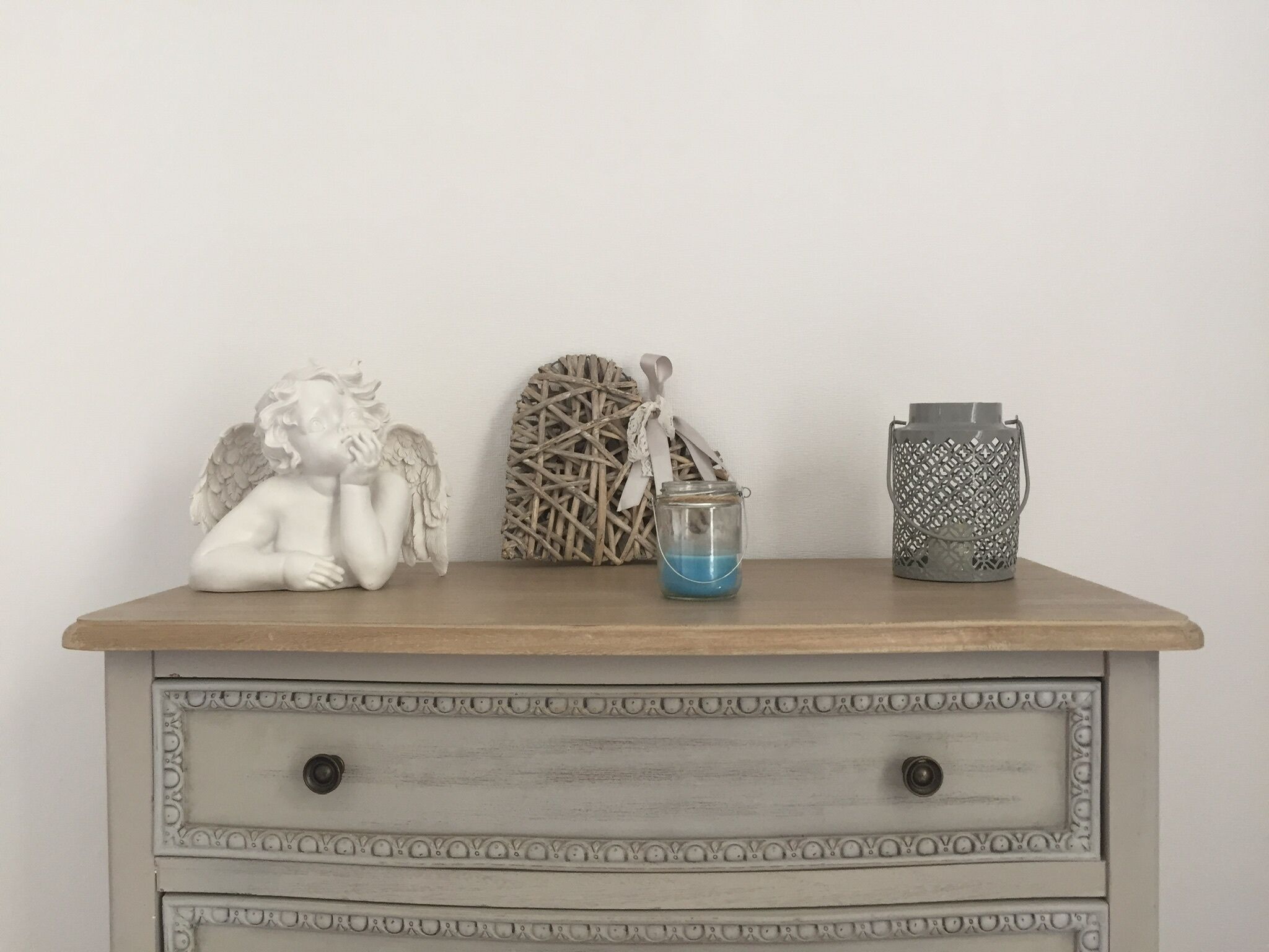 A wooden dresser with three decorative items: a white angel figurine, a woven basket with a blue candle, and a patterned metal container.