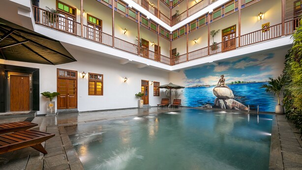 Uma área interna de piscina ampla, com um mural de uma mulher sobre uma rocha, bancos de madeira e uma varanda de dois andares com corrimãos.