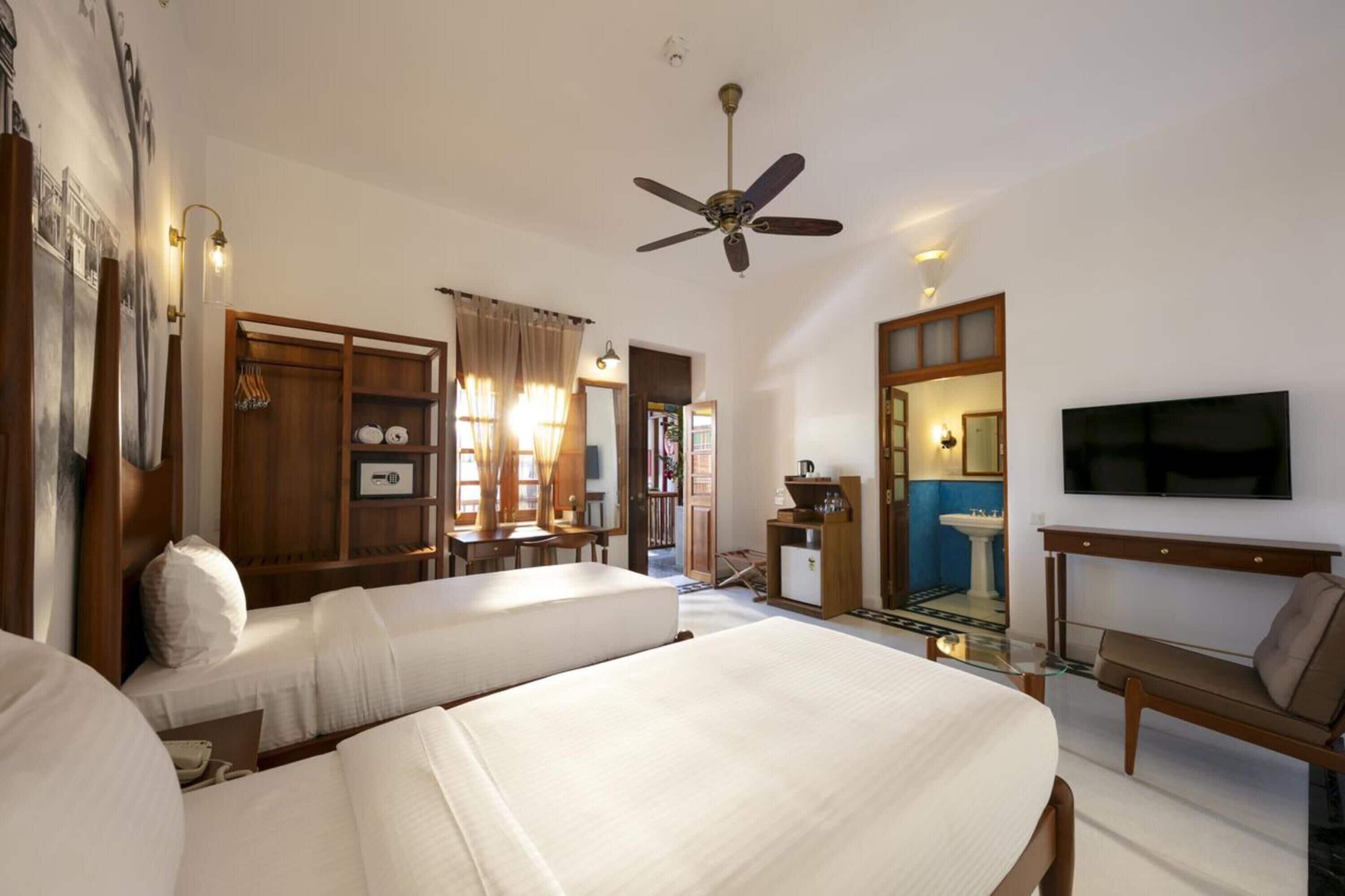 Una habitación de hotel con dos camas, un ventilador de techo, un televisor montado en la pared y una pequeña mesa con una silla.