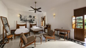 Quarto de hotel com cama, duas cadeiras, escrivaninha, ventilador de teto e um quadro na parede.
