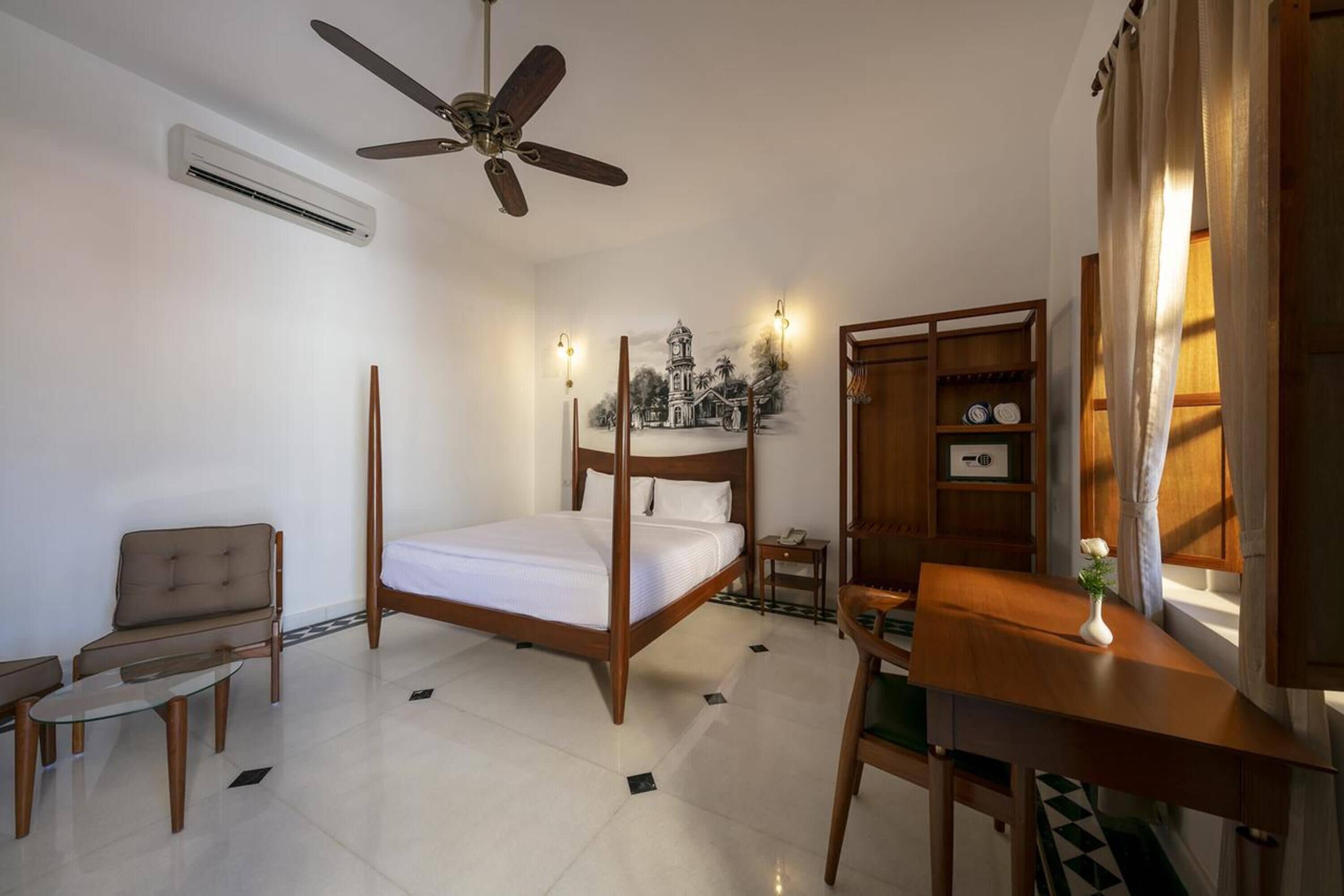 A bedroom with a four-poster bed, a ceiling fan, a wooden desk, a chair, a small table, and a wall-mounted shelf with decorative items.