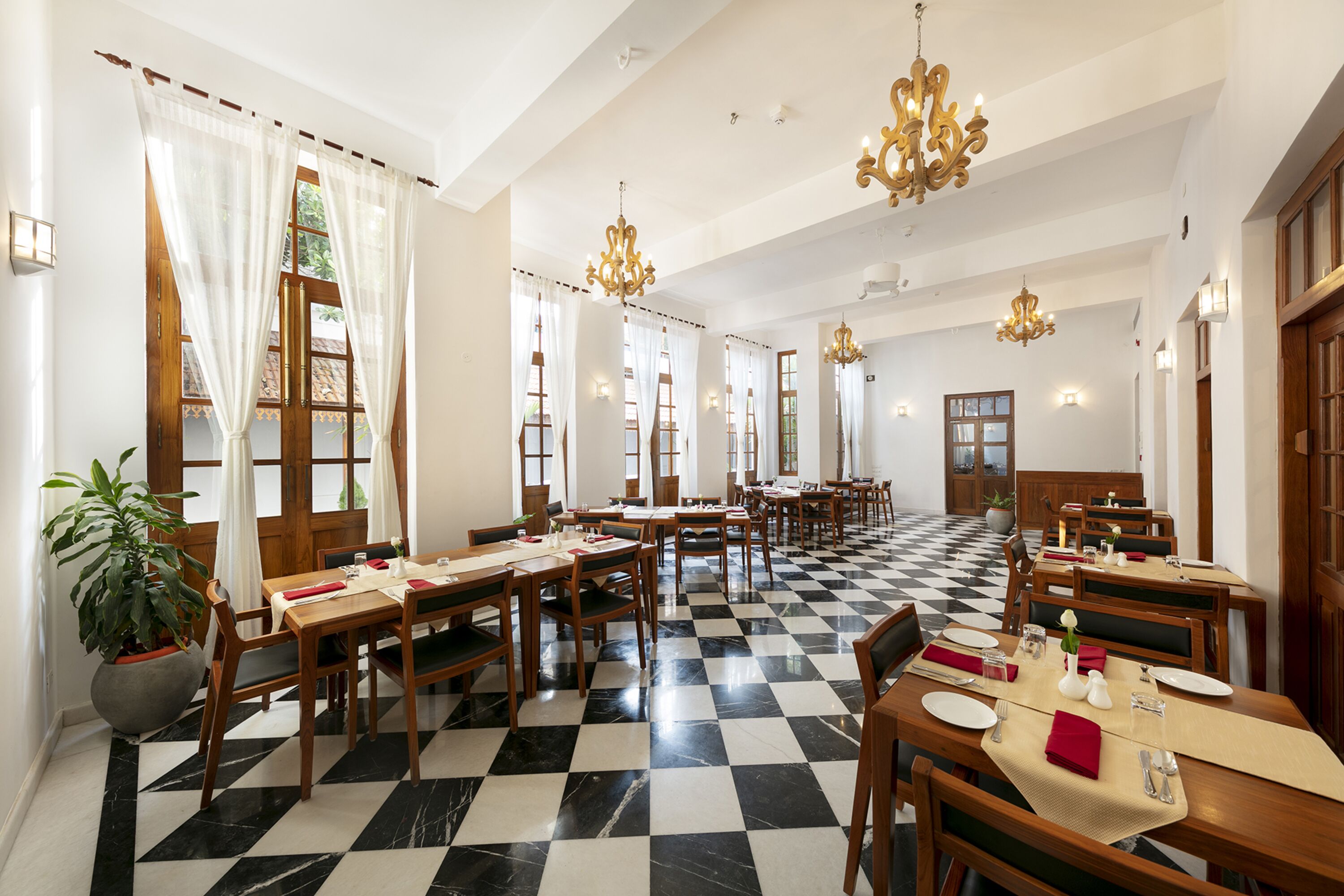 A dining area with checkered flooring, wooden tables, and chairs, featuring chandeliers and potted plants.