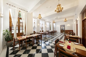A dining area with checkered flooring, wooden tables, and chairs, featuring chandeliers and potted plants. - Grand Hotel D' Europe (Puducherry)