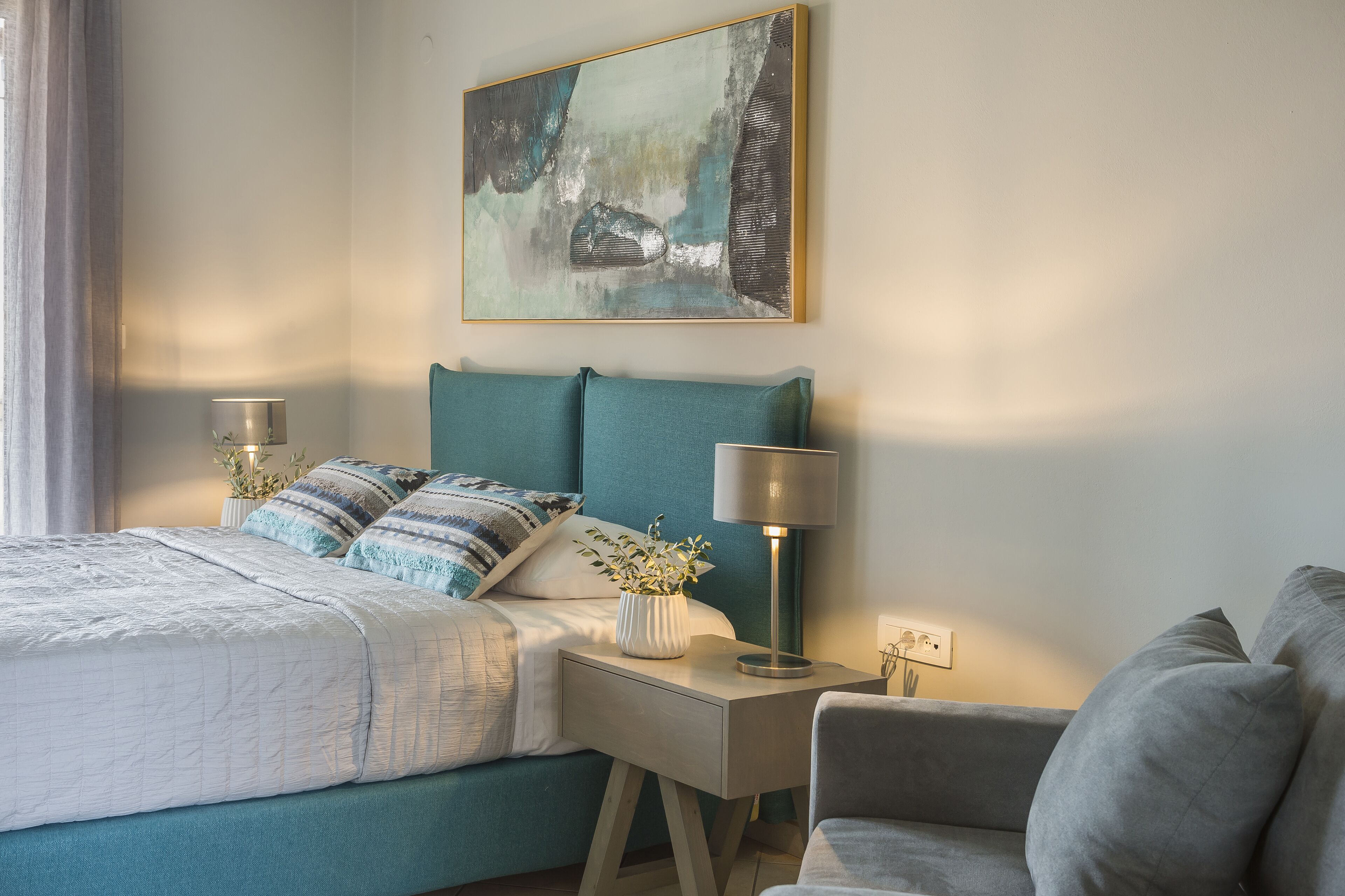 A bedroom with a bed, bedside tables, a lamp, a painting, and a plant.