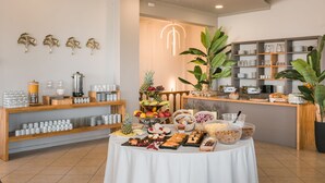A buffet setup with various food items and beverages, wooden shelves with glassware, and a large potted plant. - Makis Hotel Skala (Kefalonia)