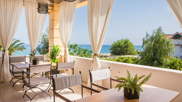 A balcony with a table and chairs, overlooking a scenic view of the sea and greenery. - Makis Hotel Skala (Kefalonia)