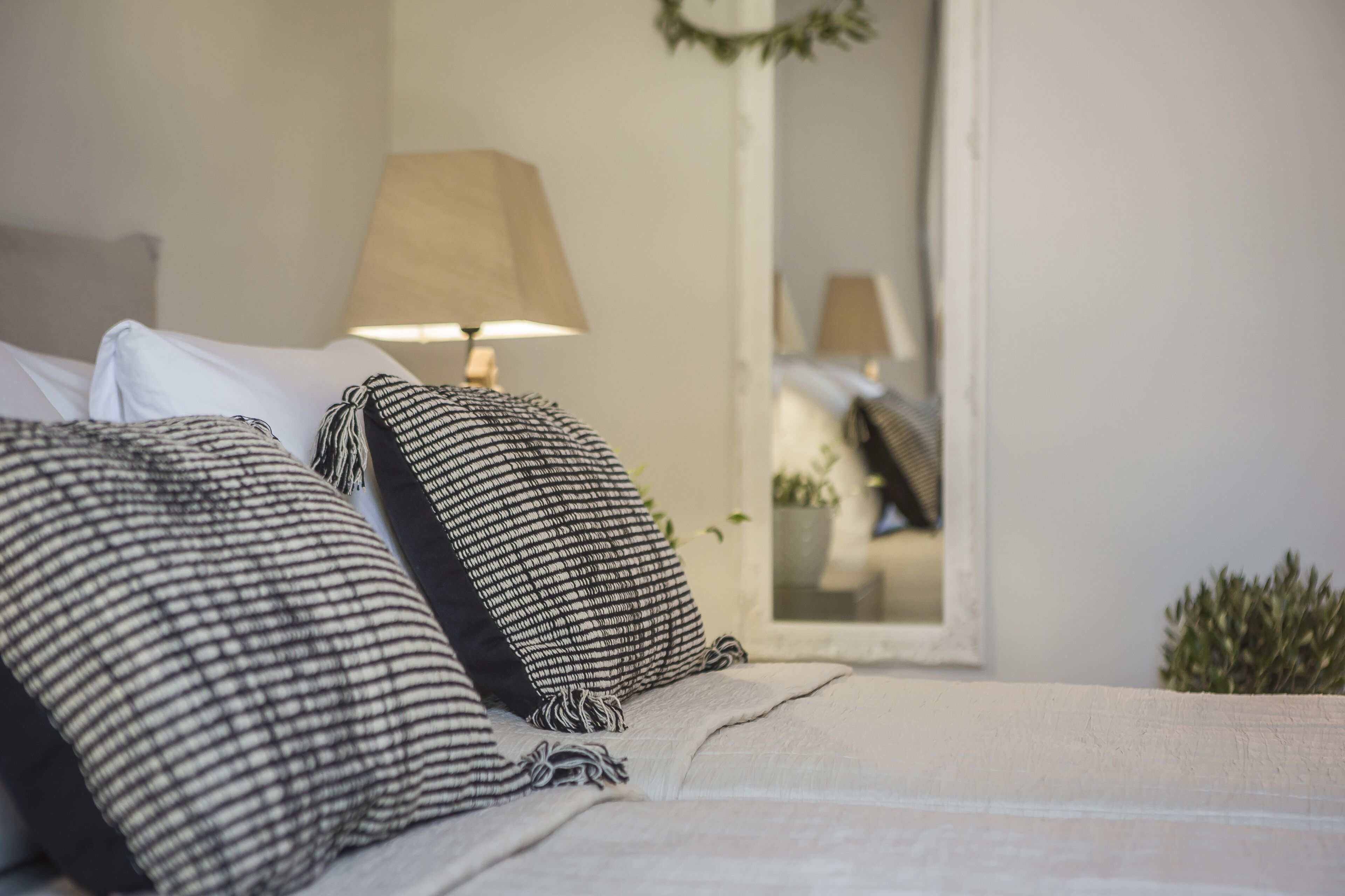 Una cama bien hecha con almohadas a rayas y una manta color beige, una lámpara en una mesita de noche y una pequeña planta junto a la cama.