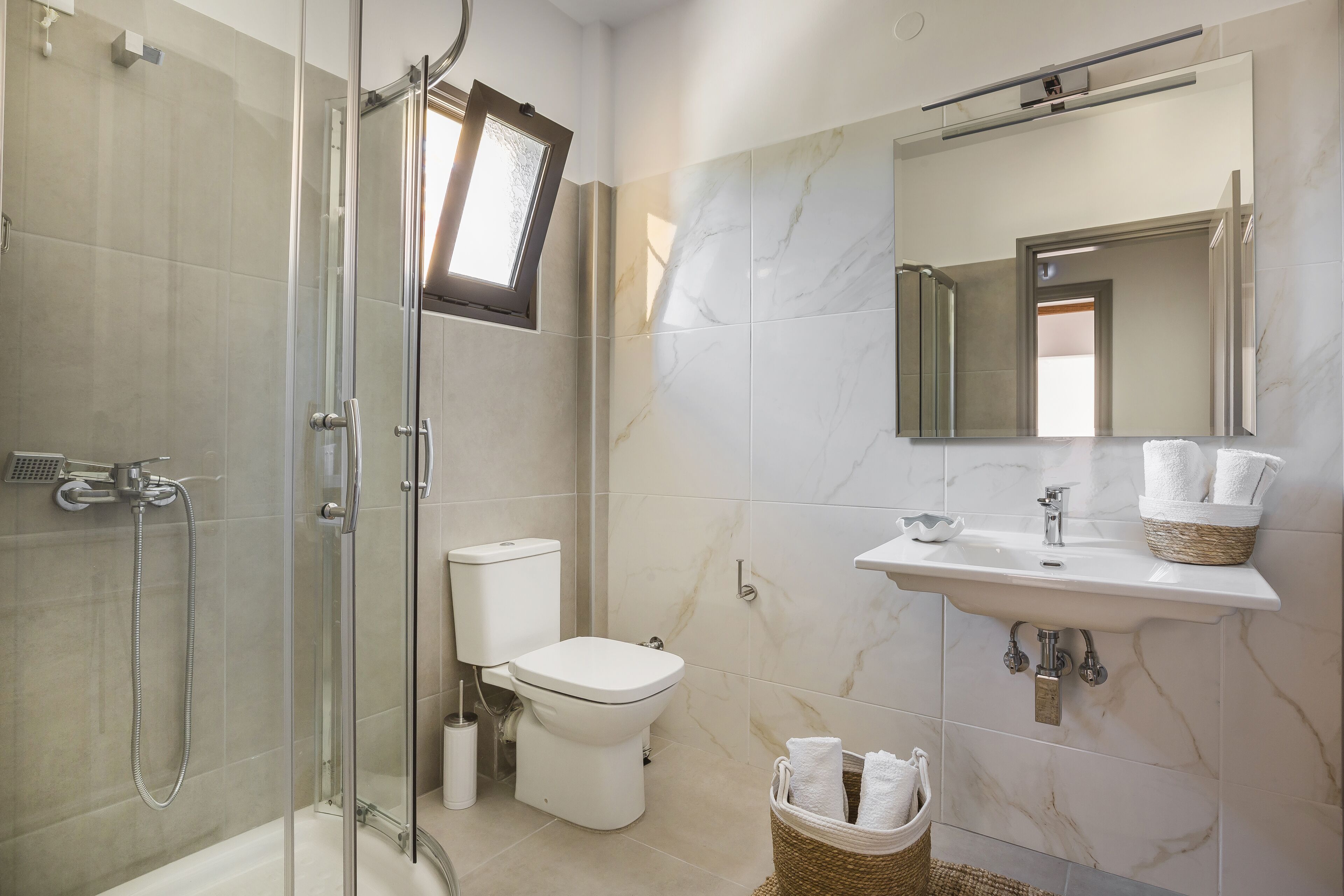 Un baño moderno con cabina de ducha de vidrio, inodoro, lavabo con encimera de mármol y espejo sobre el lavabo.