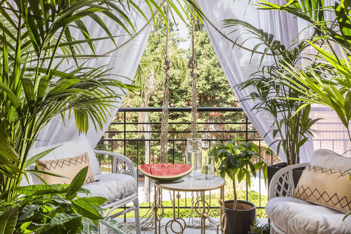 A cozy outdoor seating area with a white wicker chair, a round table with a watermelon, a glass of water, and a potted plant.