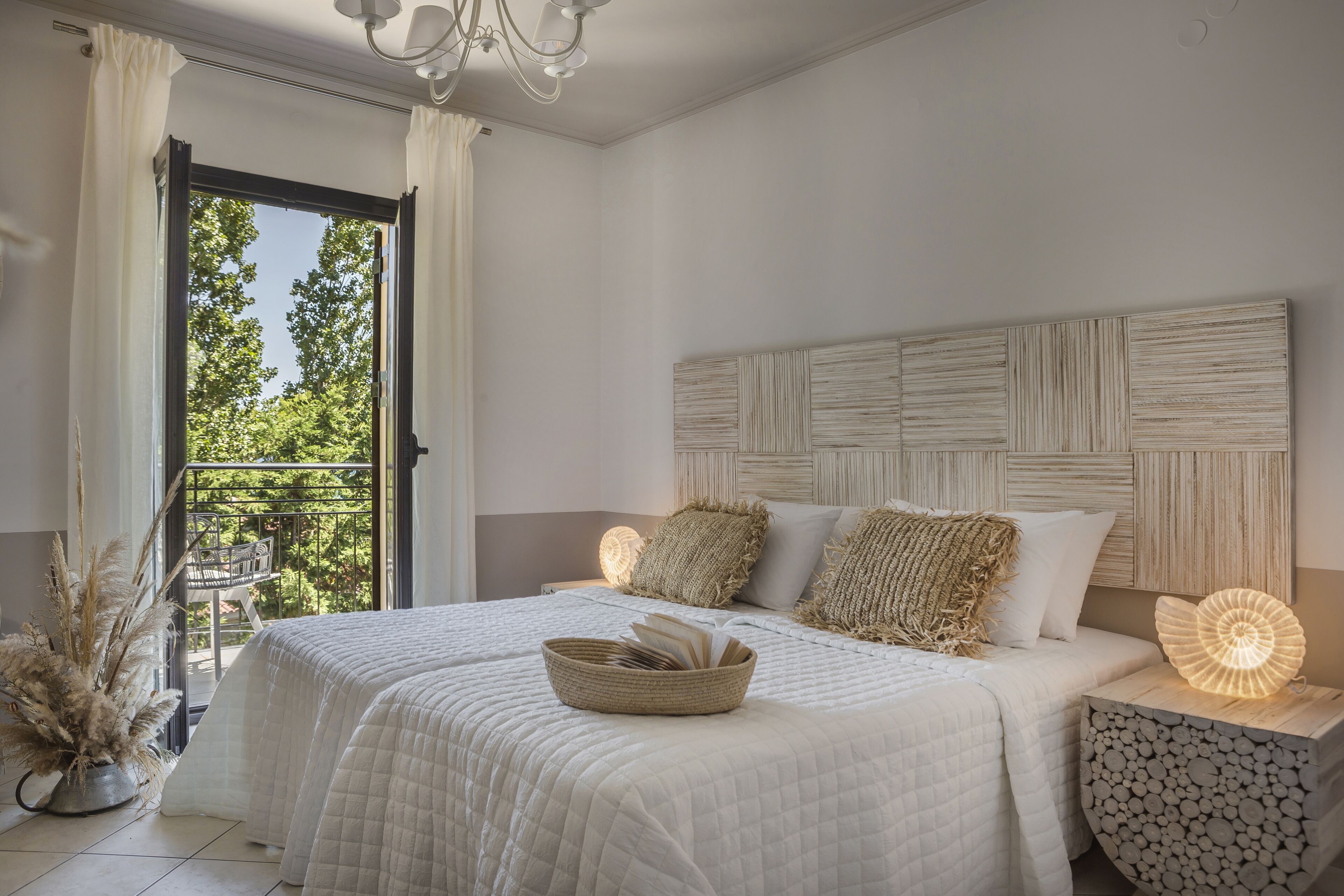 Une chambre avec un grand lit, une table de chevet avec une lampe, une vue sur l’extérieur par une fenêtre et un panier décoratif posé sur le lit.