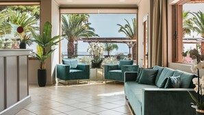 A spacious living area with a green sofa set, a potted plant, and a view of palm trees through large windows. - Makis Hotel Skala (Kefalonia)