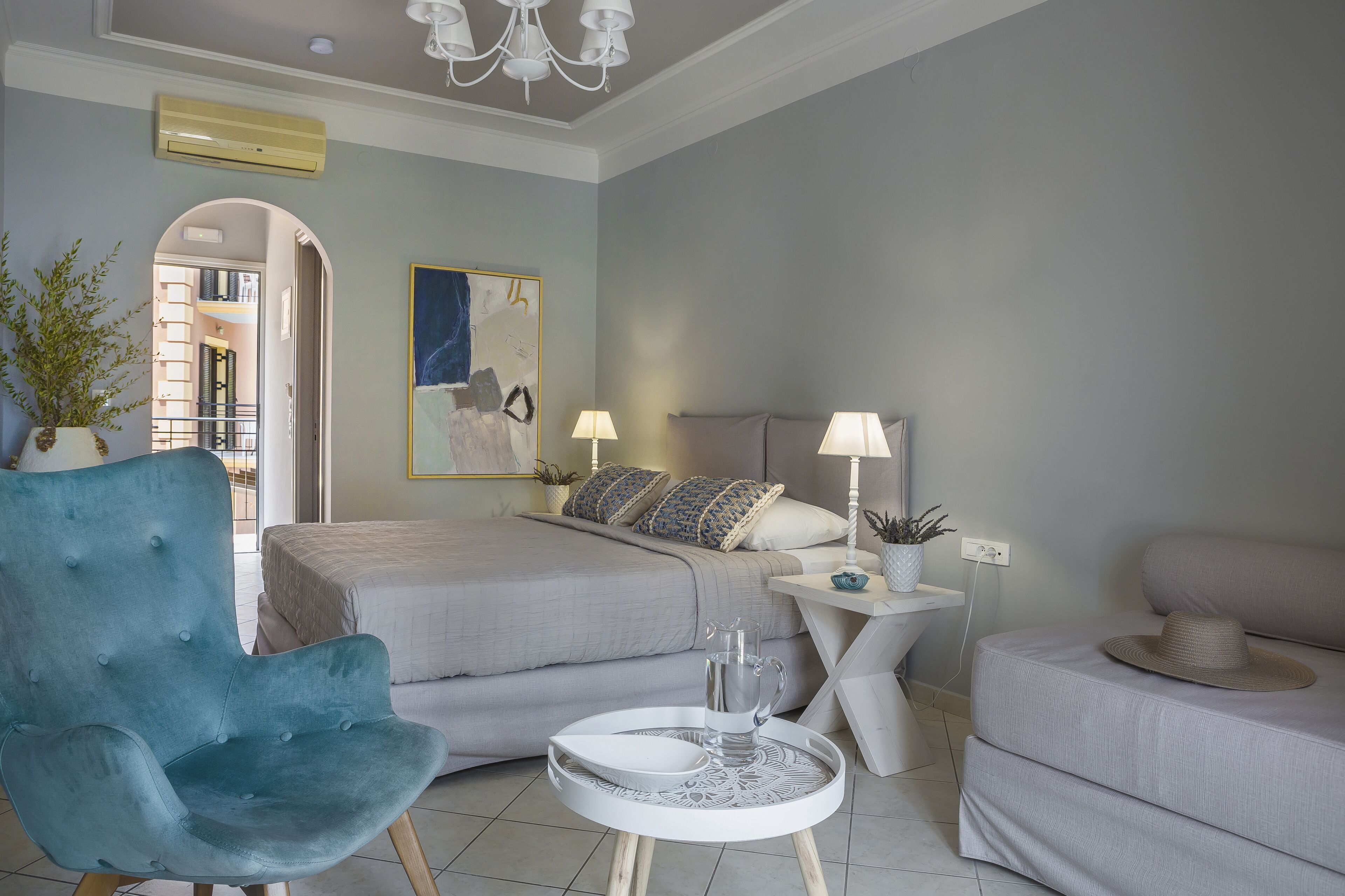 A modern bedroom with a bed, a blue armchair, a small round table with a pitcher, and a bedside lamp.