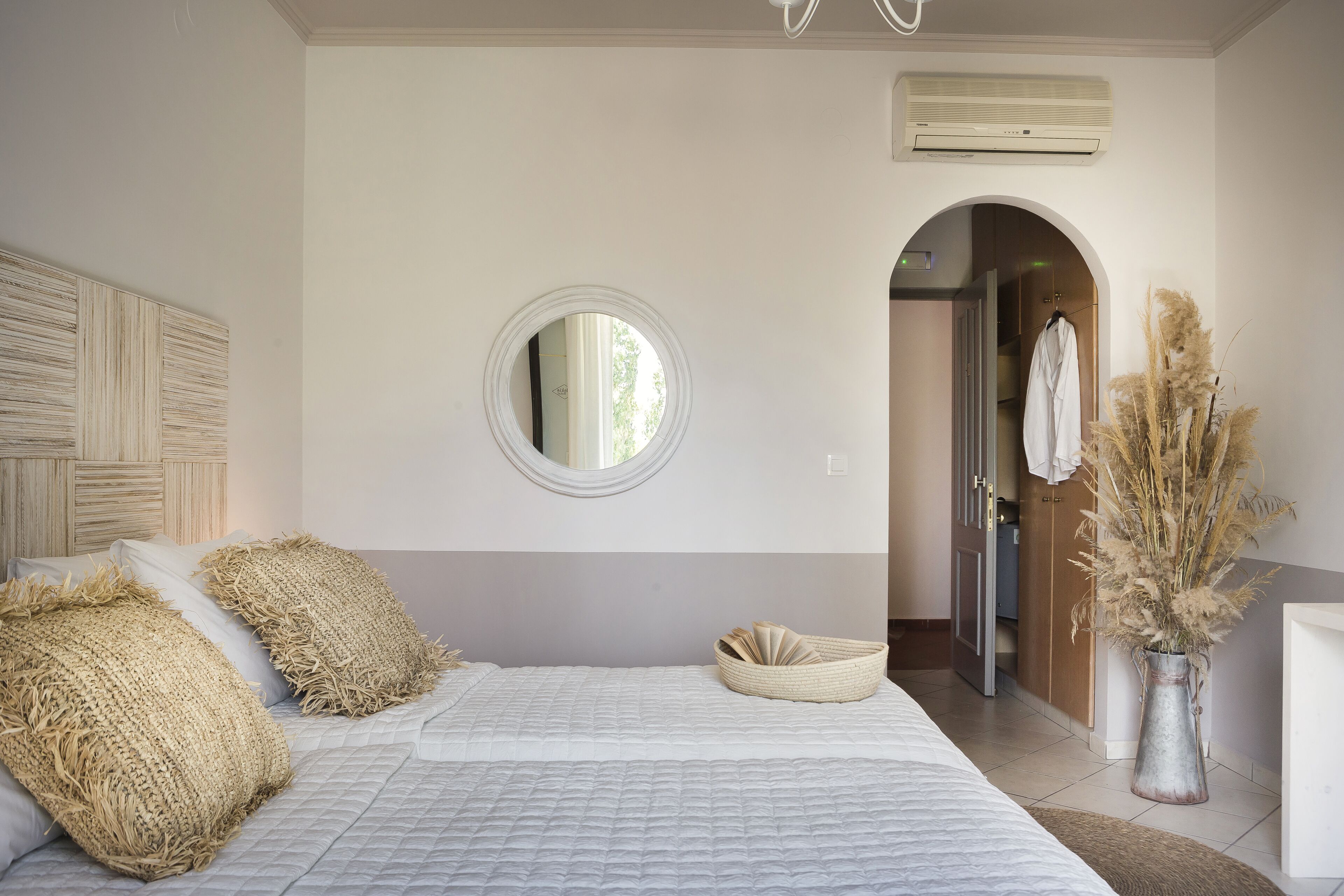Un dormitorio con cama, almohadas, un espejo redondo, una mesita de noche, un jarrón con plumas de pampa y una puerta que da a otra habitación.