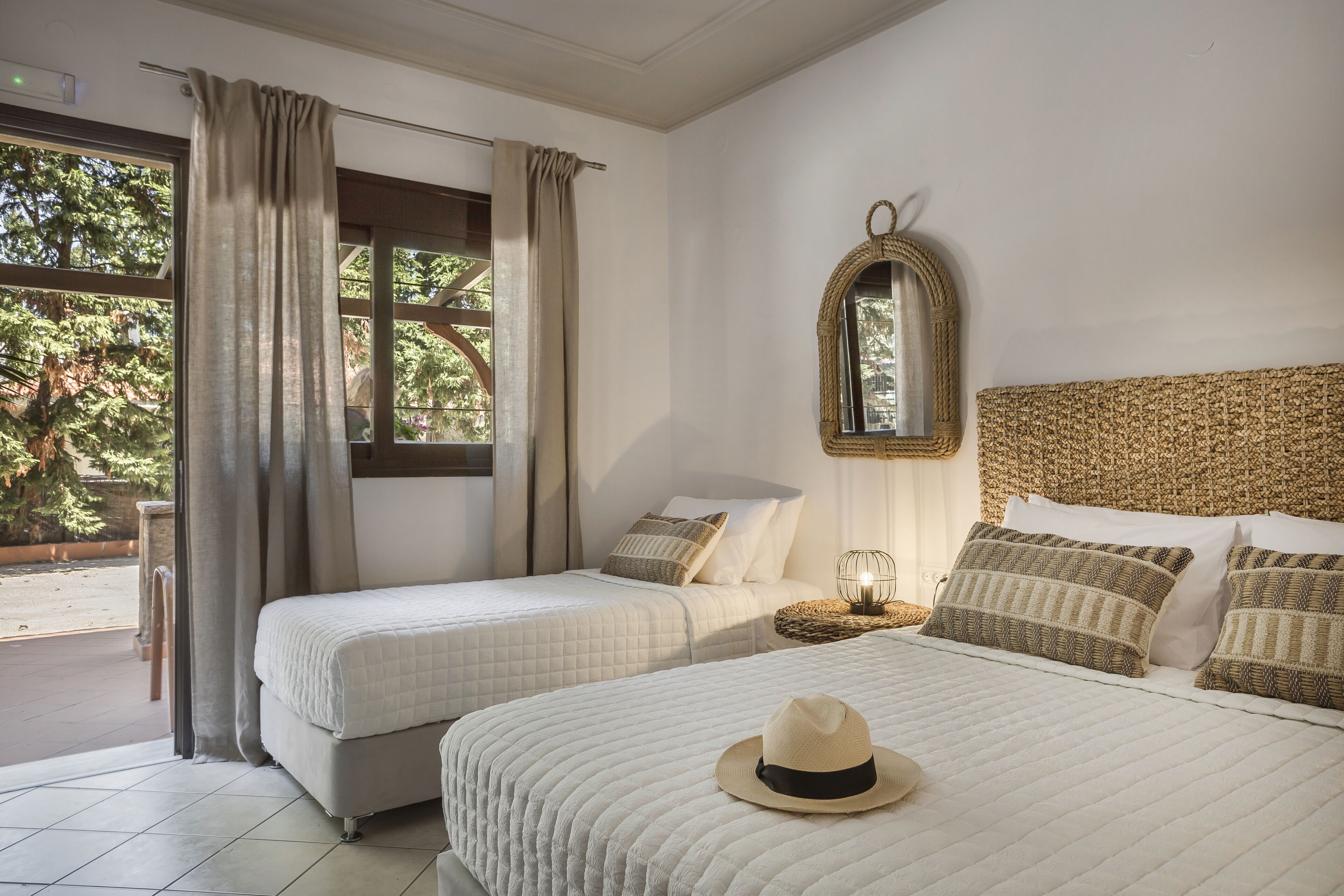 A hotel room with two beds, a large window with curtains, a woven headboard, and a mirror.