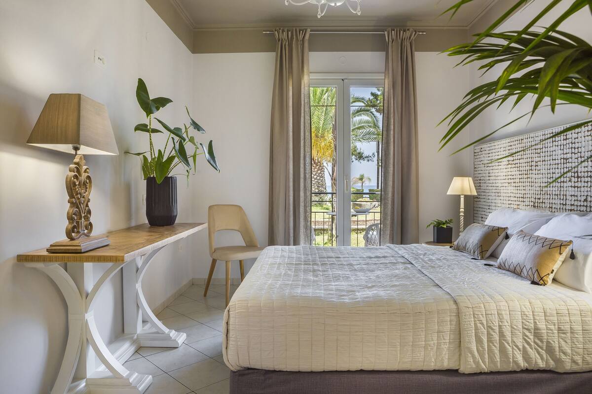 A hotel room with a bed, a desk, a chair, a potted plant, and a view of the outdoors.